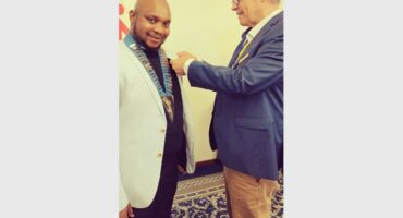 A black male wearing a white blazer and black shirt smiles at the camera while a white male pins something on his jacket.