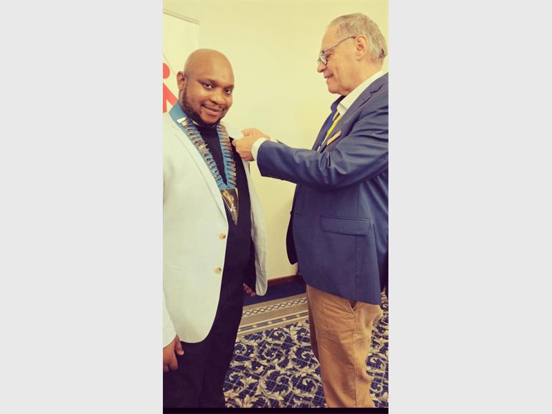 A black male wearing a white blazer and black shirt smiles at the camera while a white male pins something on his jacket.