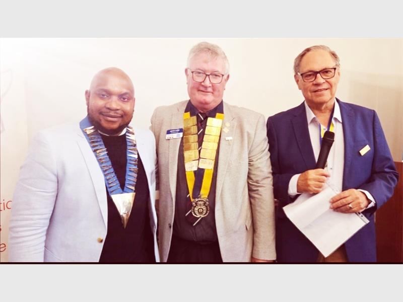 Three men in a row, two wearing rotary crests around their necks and the third holding a piece of paper and microphone