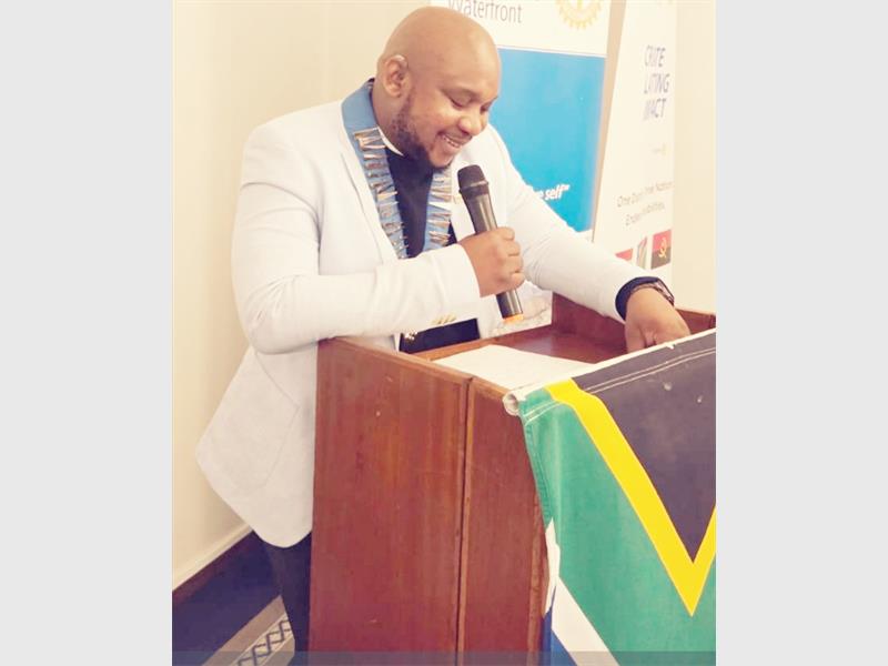 A black male wearing a white blazer with a blue sash around his neck holding a microphone behind a podium adorned with the SA flag