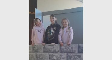 Three young girls standing behind a wall used as a prop on a stage where they are rehearsing.