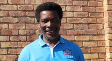 Black man wearing a light blue ACDP shirt standing in front of a brick wall