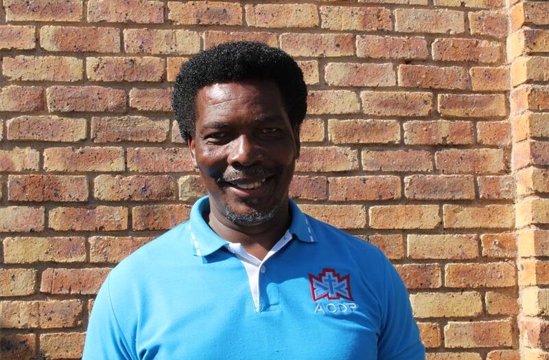 Black man wearing a light blue ACDP shirt standing in front of a brick wall