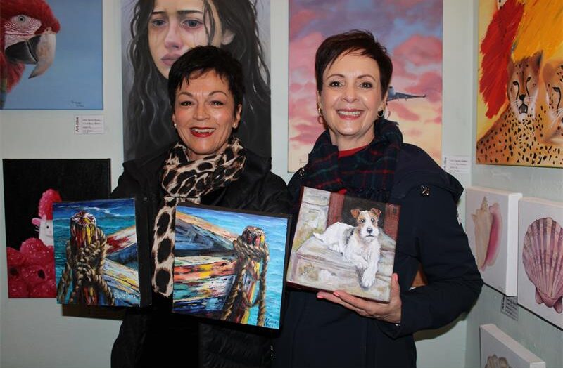 Two women holding canvasses stand in front of a wall adorned with art