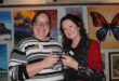 Two women hold their glasses together as they stand in front of a wall adorned with art