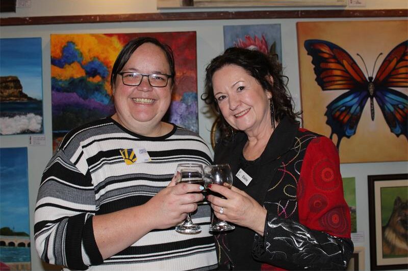 Two women hold their glasses together as they stand in front of a wall adorned with art
