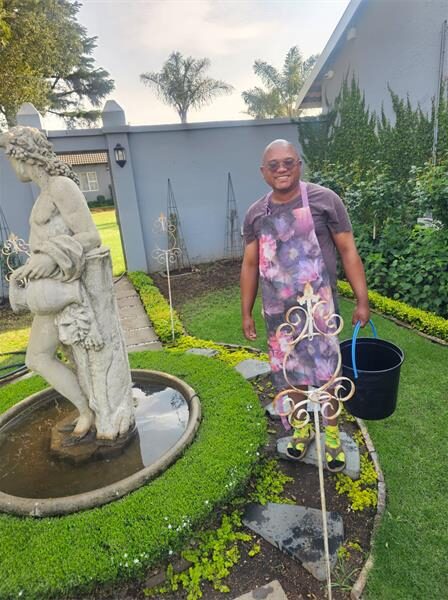 Coloured man in a floral apron holding a black bucket standing next to a water feature