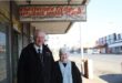 An elderly man and women stand next to a street under a weather-worn sign