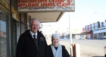 An elderly man and women stand next to a street under a weather-worn sign