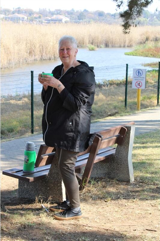 Elderly woman stands at a bench, holding a cup of coffee with a dam in the background