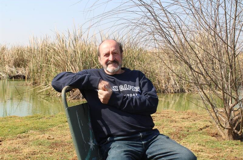 Elderly man sitting on a bench with reeds in the background while he smiles at the camera