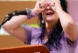 A woman behind a podium with a microphone on it holding her hands up to her eyes like binoculars.