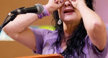 A woman behind a podium with a microphone on it holding her hands up to her eyes like binoculars.