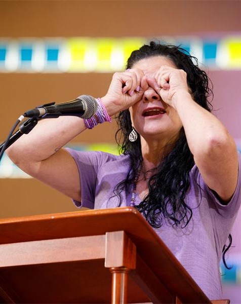 A woman behind a podium with a microphone on it holding her hands up to her eyes like binoculars.