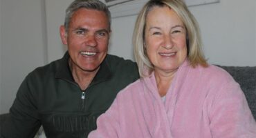 Man smiles as he sits next to a woman wearing a pink robe