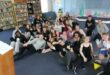 A big group of men and women sitting on a grey carpeted floor in various poses.