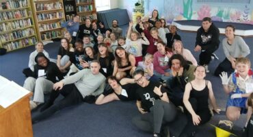 A big group of men and women sitting on a grey carpeted floor in various poses.