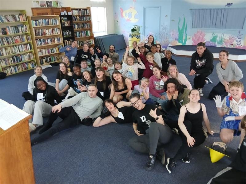 A big group of men and women sitting on a grey carpeted floor in various poses.