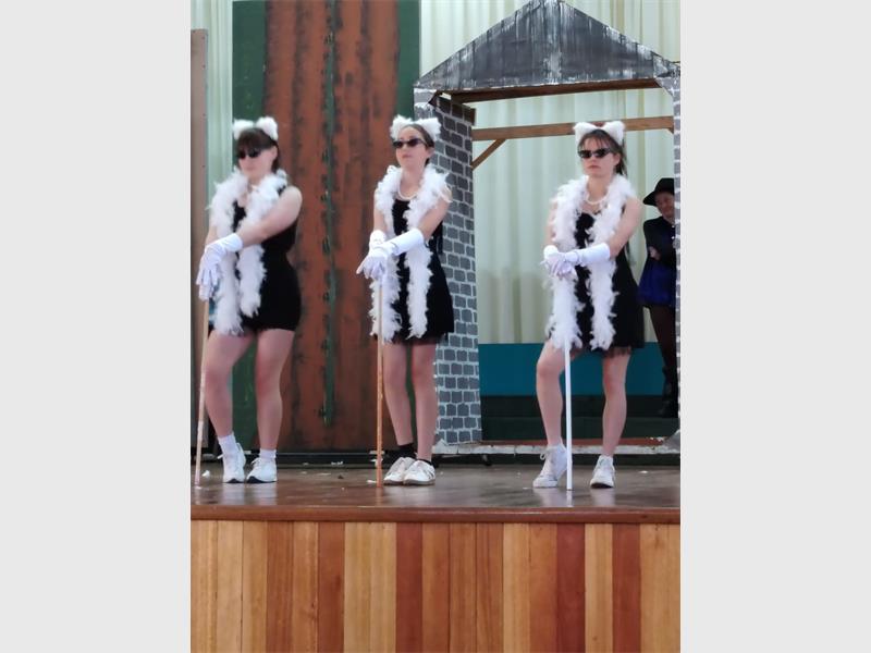 Three young girls on stage dressed in short black dresses, wearing dark sunglasses with white boas on and walking sticks.