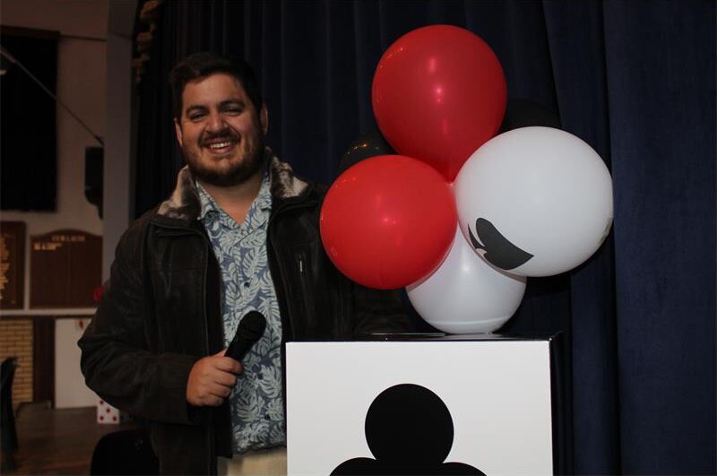 White man with a beard holds a microphone while posing next to red and white balloons