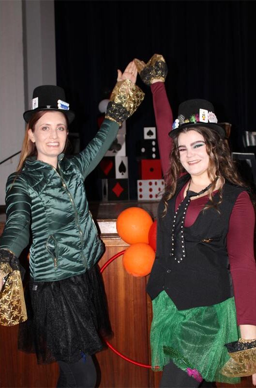 Two white women, wearing top hats hold their hands up in the air