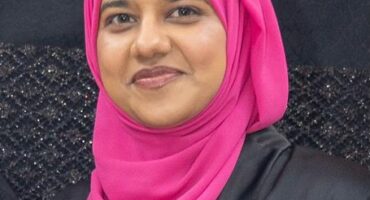 A head and shoulders photo of an Indian woman wearing a black dress and pink headscarf.