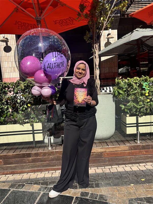 An Indian woman dressed in black with a beige headscarf holding a book and a bunch of purple and pink balloons.