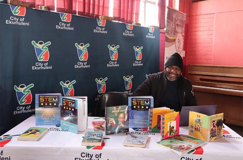 A man sitting by a book stall wearing a black jacket and a black hat