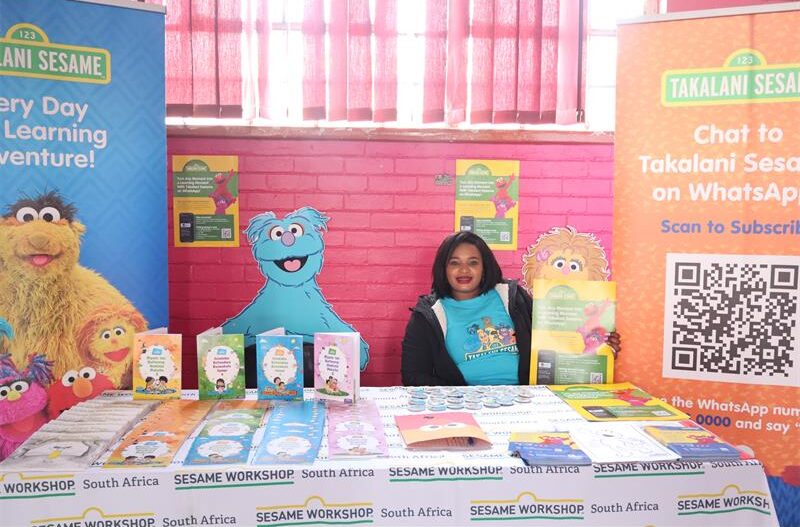 Lady sitting by a book stall wearing a blue top and a black jacket, holding a pamphlet 