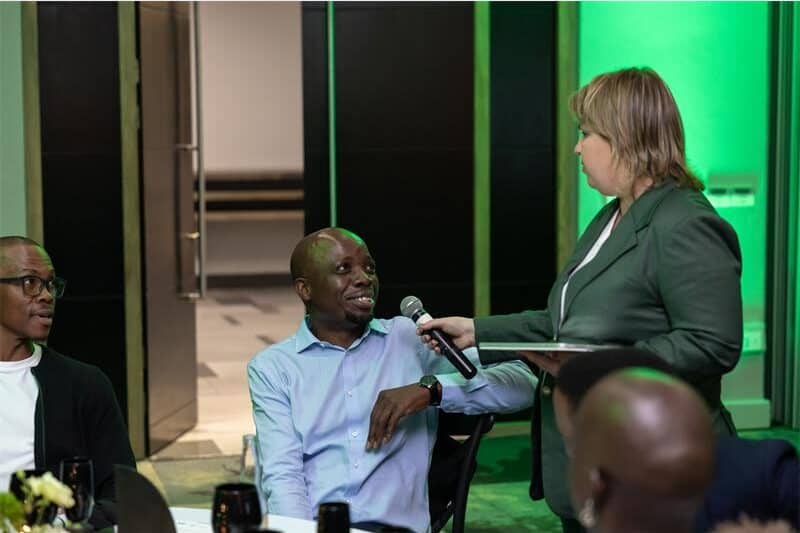 Two black males seated at a table while a white woman stands alongside one of them and holds a microphone out to him.