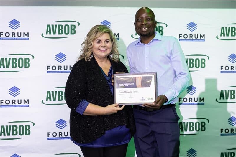 A white woman and a black man on stage next to each other, holding a certificate in front of a banner with FCJ and AVBOB logos.