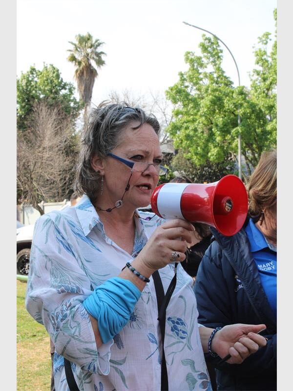 A white woman with grey hair and glasses holding a megaphone with people in the background.