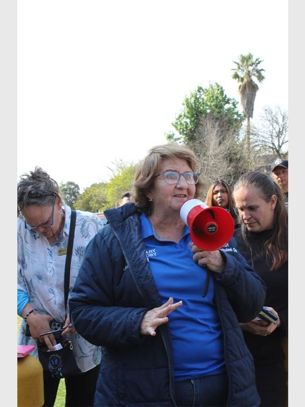 An elderly woman dressed in a blue jacket holding a megaphone with people in the background.