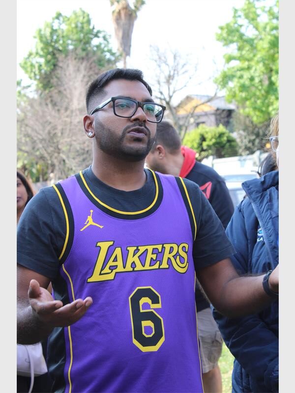 An Indian male dressed in a purple shirt with his arms gesturing while speaking, and wearing glasses.