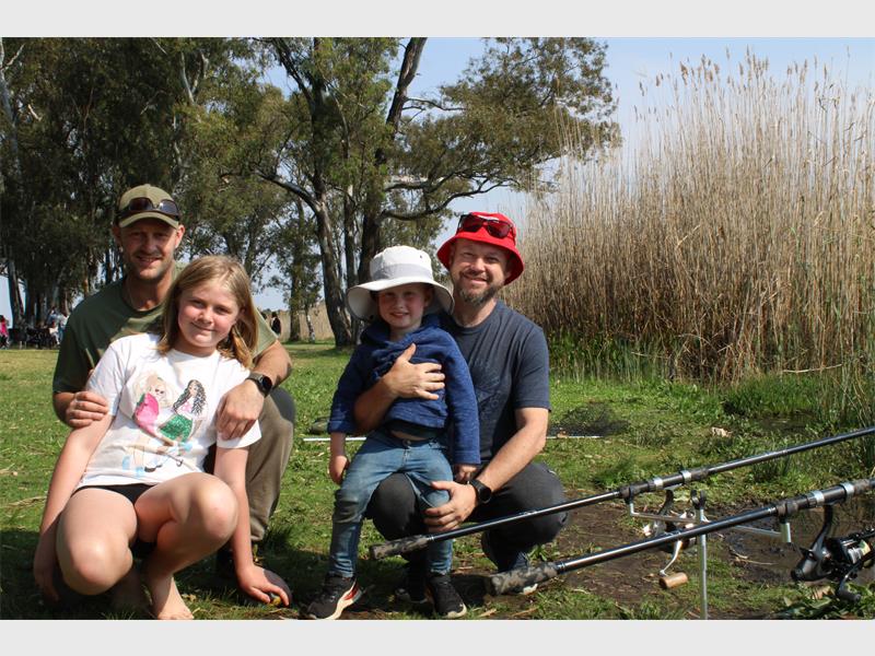 Two men with a young girl and a young boy on the banks of a dam with fishing rods.