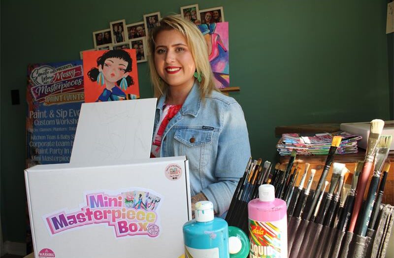 Blonde woman holding a white pox with writing on it and art works behind her