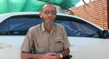 Bearded white man sitting in a wheelchair in front of a white car