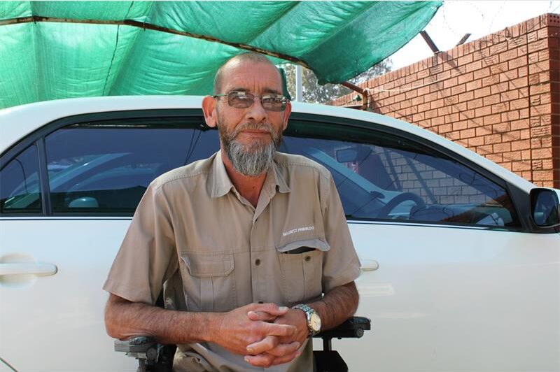Bearded white man sitting in a wheelchair in front of a white car