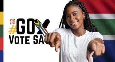 A young black female with dreadlocks pointing into the camera wearing a white T-shirt with a cross and the words go vote SA and the SA flag colours next to her.