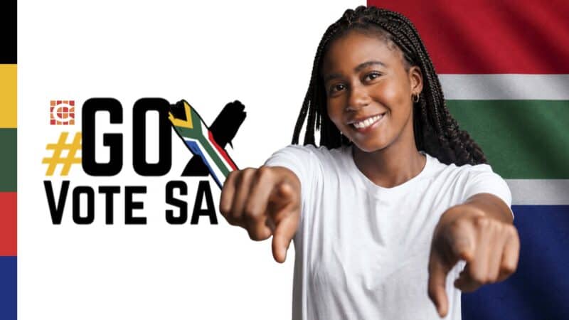 A young black female with dreadlocks pointing into the camera wearing a white T-shirt with a cross and the words go vote SA and the SA flag colours next to her.