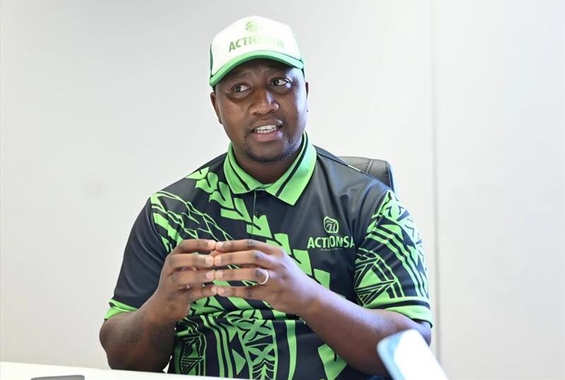 A black man dressed in a green and black cap and shirt with his hands clasped together.