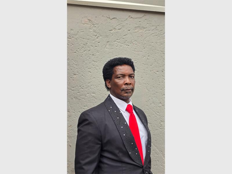 A black male standing in front of a wall wearing a grey suit and red tie.