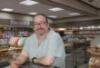 White man with long grey hair and wearing glasses leaning on a counter with baked goods in the background