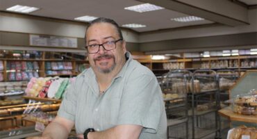 White man with long grey hair and wearing glasses leaning on a counter with baked goods in the background