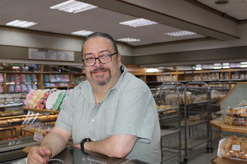 White man with long grey hair and wearing glasses leaning on a counter with baked goods in the background