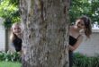 Two teenage girls wearing black looking out from behind a tree