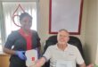 Swart vrou met blou handskoene staan langs 'n wit man met 'n plakaat wat sê hy skenk sy 100ste eenheid van bloed