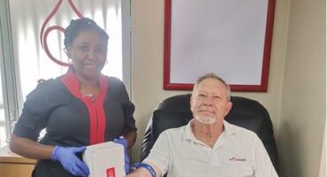 Swart vrou met blou handskoene staan langs 'n wit man met 'n plakaat wat sê hy skenk sy 100ste eenheid van bloed