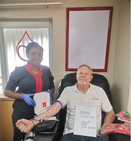 Swart vrou met blou handskoene staan langs 'n wit man met 'n plakaat wat sê hy skenk sy 100ste eenheid van bloed