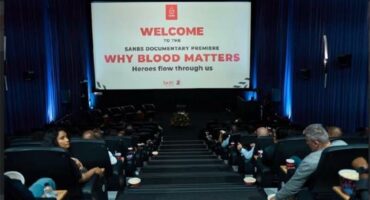 A movie theatre photographed from the door towards the screen showing people in seats and the message welcome to why blood matters on the screen.